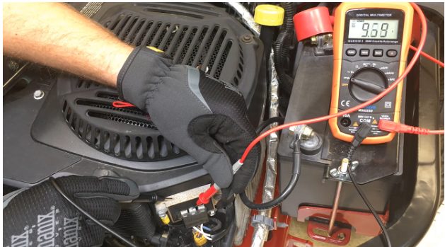A mechanic with black gloves testing battery with multimeter at 9.69 reading