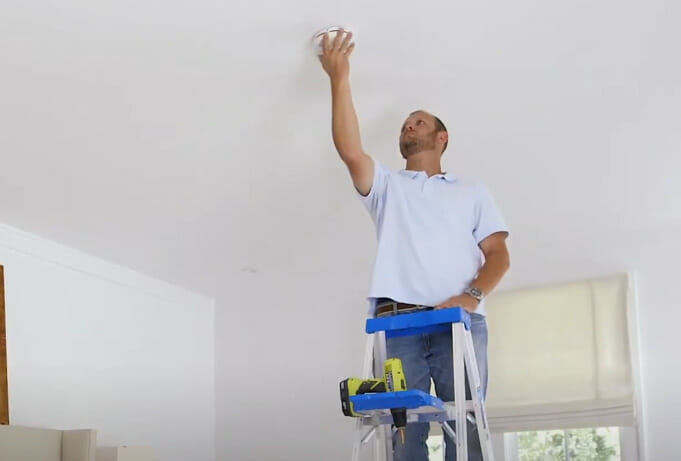 A person with a ladder reaching out to a smoke detector at the ceiling