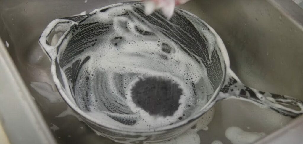 A person washing an iron cast pan with dishwashing soap at a kitchen sink
