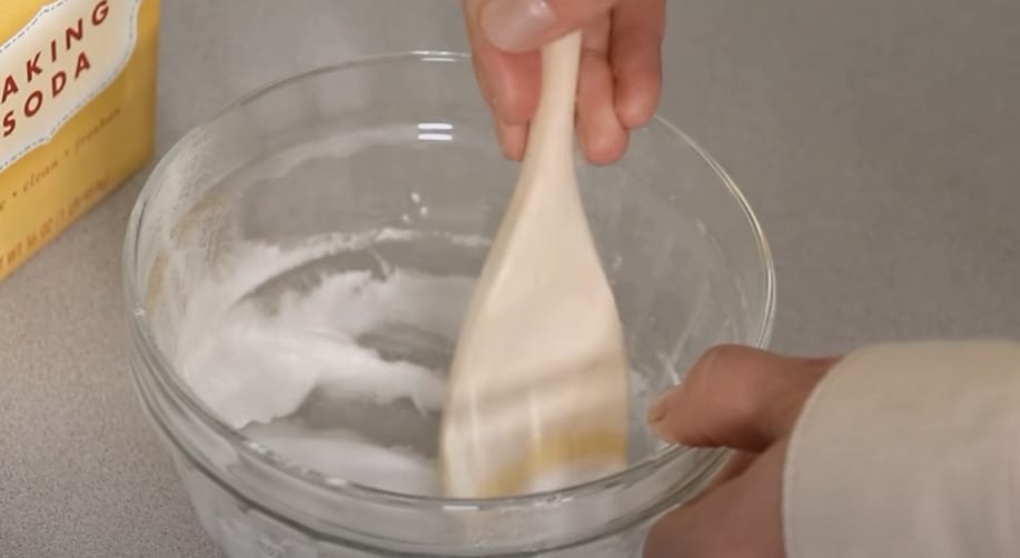 A person using a wooden spoon to stir baking soda and water in a glass bowl