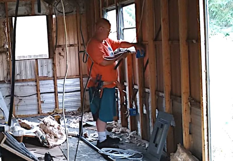 A man in an orange shirt is working on a house, specifically focusing on how to run electrical wire