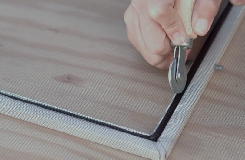 A person performing home repairs, using pliers as a guide while cutting a screen on a window