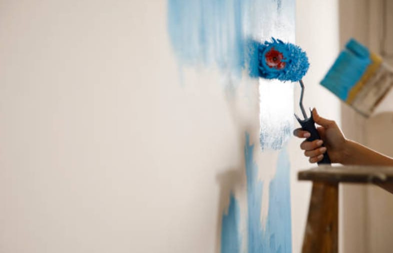 A woman is repainting the wall in blue using a roller