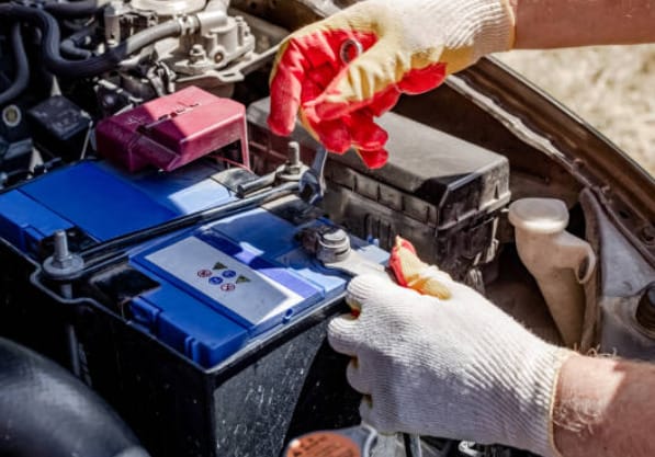A man wearing a yellow red gloves is testing a car battery using a multimeter