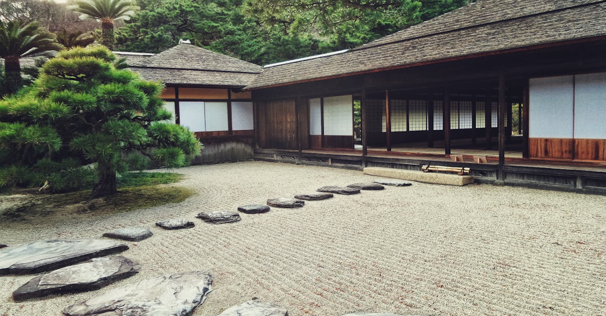 A traditional Japanese Zen garden with raked gravel, stepping stones, a small tree, and a wooden building in the background.