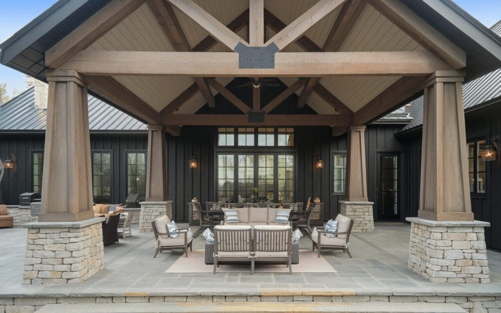 An outdoor patio with wooden beams, stone pillars, and a seating area with cushions. This Modern Farmhouse retreat connects to a house with large glass windows and dark-colored exterior walls.
