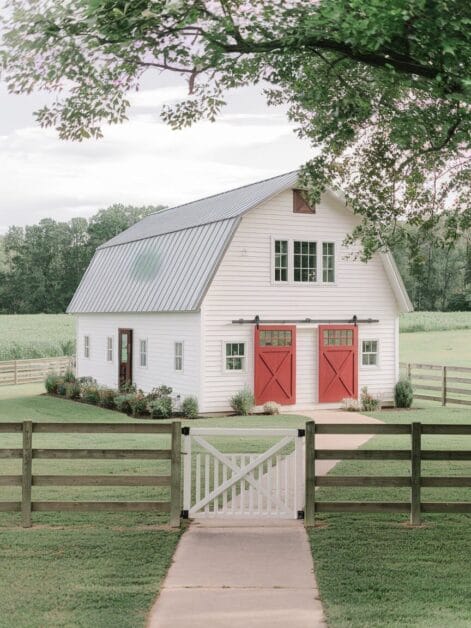 A white barn with a gray metal roof and two red doors sits in a fenced yard surrounded by greenery and trees—perfect for Barndominiums enthusiasts seeking their dream home under $120K.
