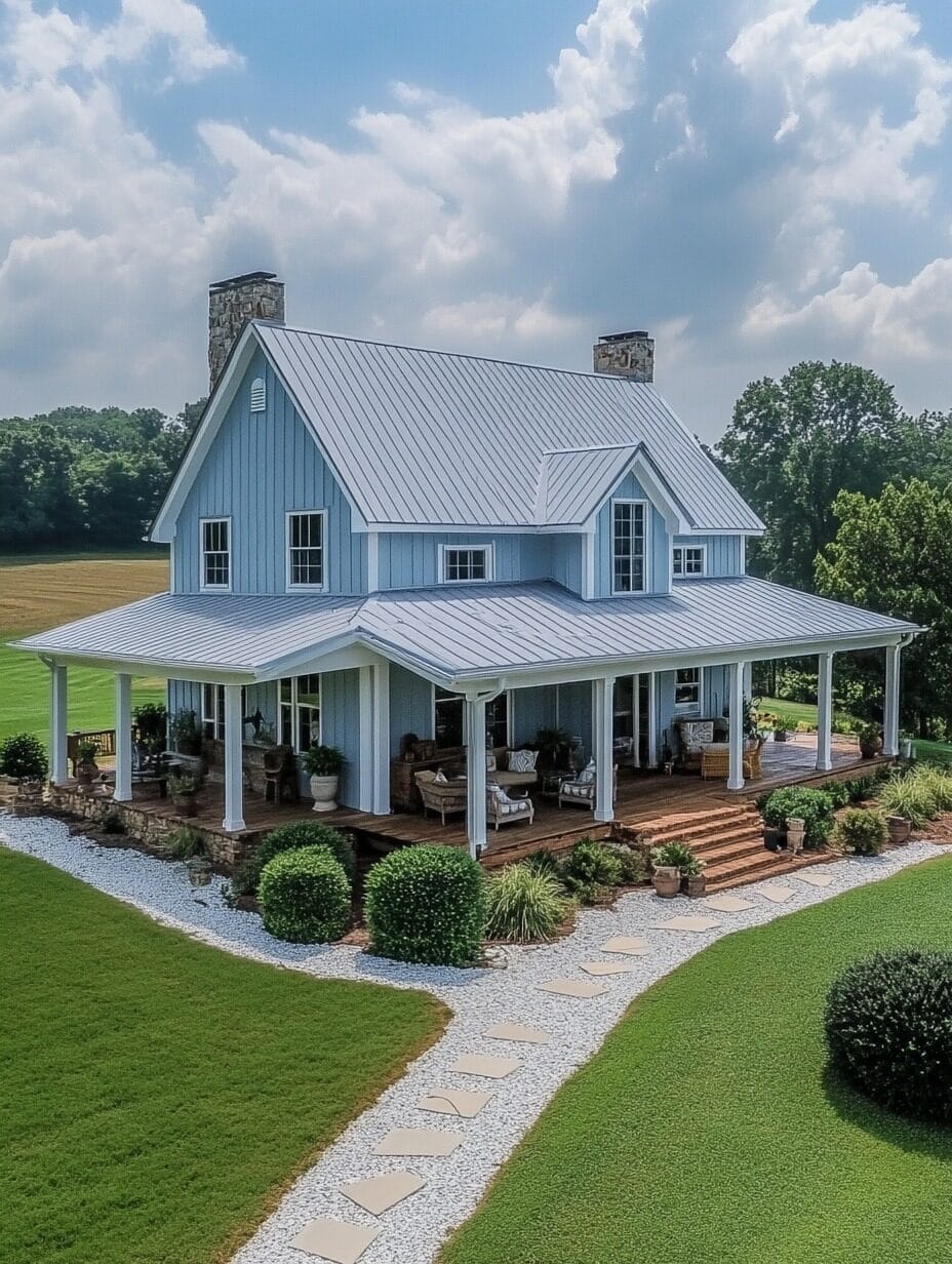 A two-story blue farmhouse with a metal roof, stone chimneys, and a large wraparound porch showcases charming barndominium designs, surrounded by green lawn and landscaping under a partly cloudy sky.