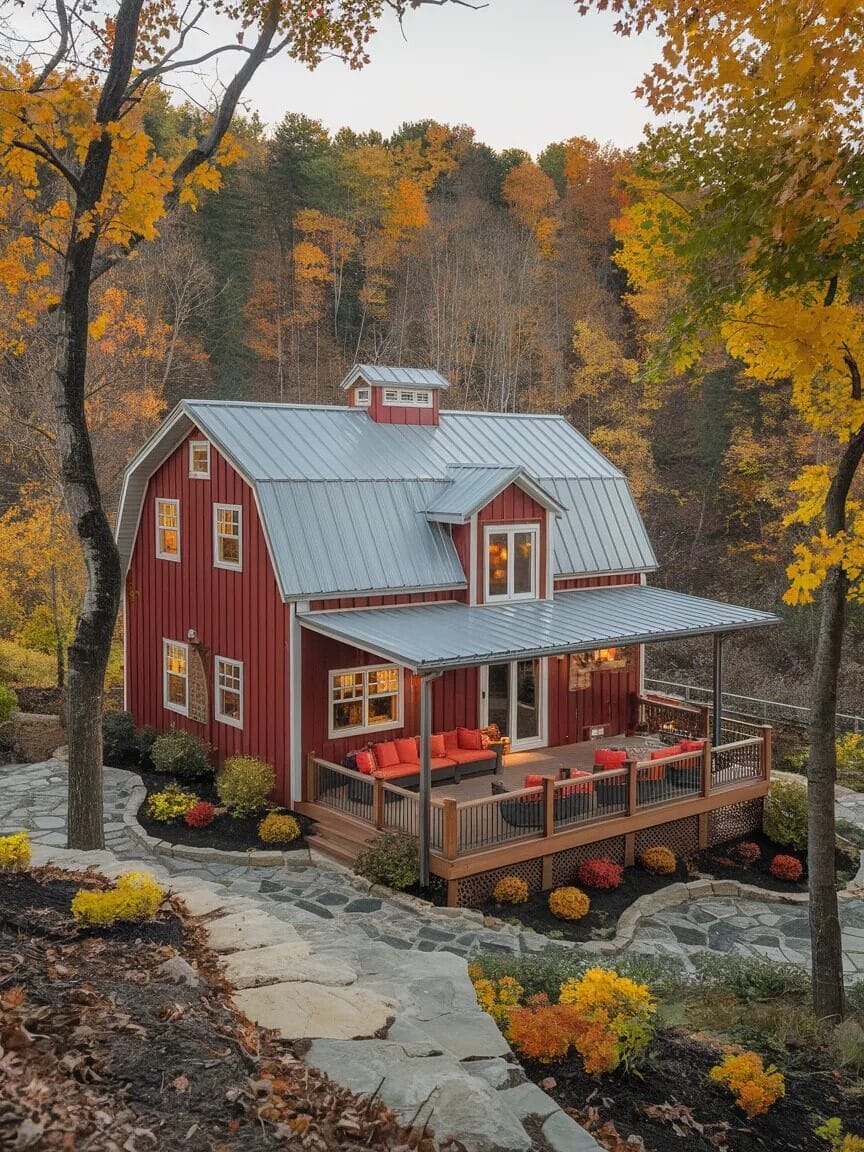 A red barn-style Barndominium with a metal roof and wraparound porch sits among autumn trees with colorful foliage and landscaped stone pathways—the perfect dream home for any budget.