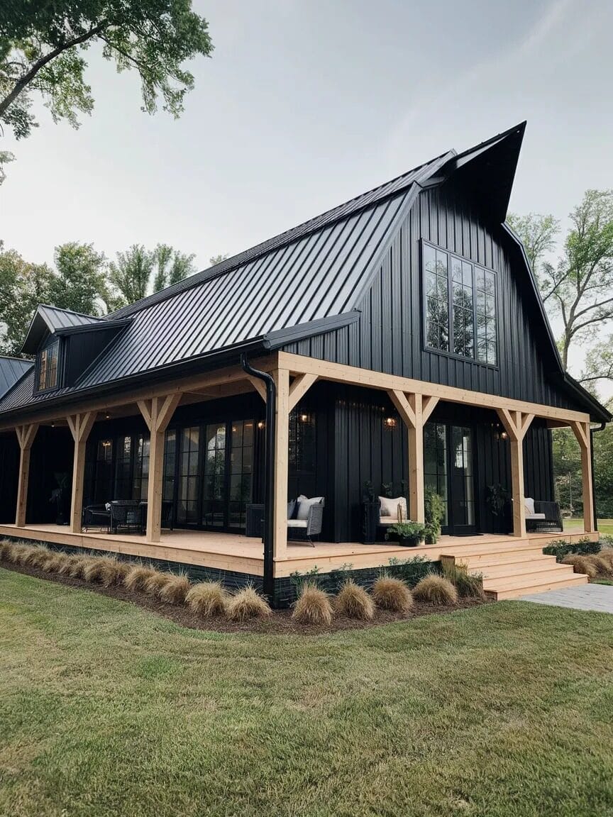 A modern black barn-style house with a metal roof, large windows, wooden columns, and a wraparound porch offers the charm of stunning barndominiums, surrounded by grass and landscaping.