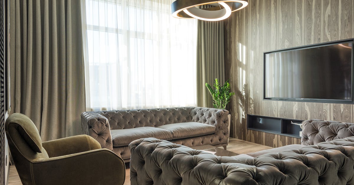 Modern living room with tufted sofas, an armchair, a large wall-mounted TV, tall curtains, a circular ceiling light, and a potted plant near the window.