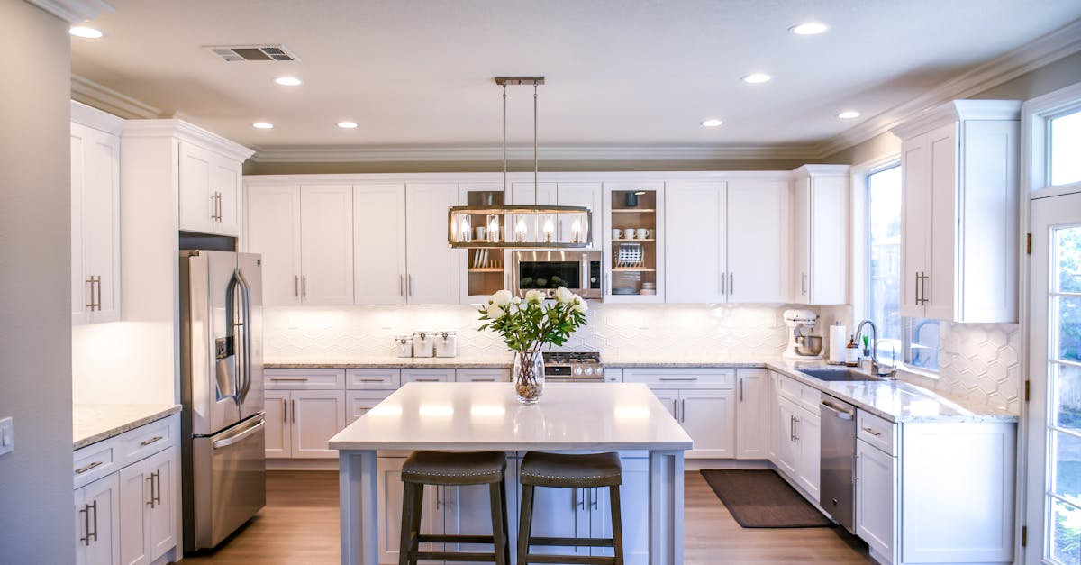 Modern kitchen with white cabinets, stainless steel appliances, center island with two stools, vase of flowers, and large windows letting in natural light.