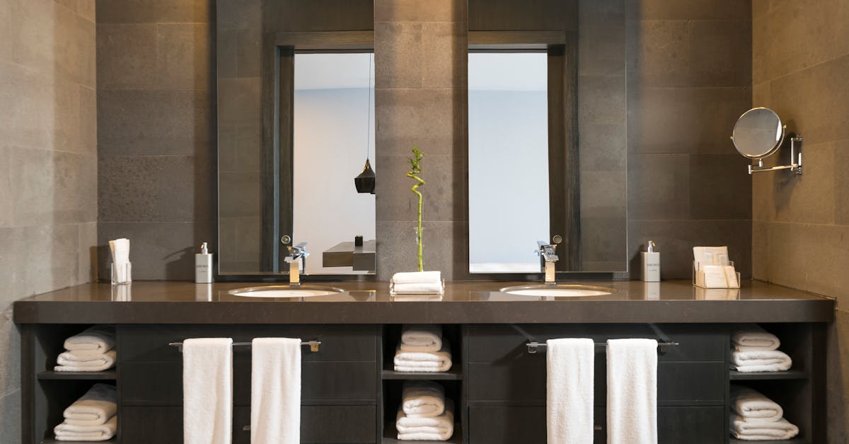 Modern bathroom with two sinks, mirrors, wall-mounted faucets, stacked white towels, and a single bamboo stalk in a vase on the dark countertop.