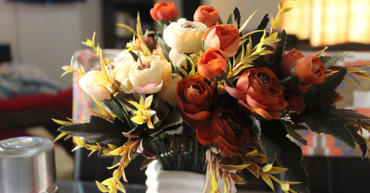 A close-up of a bouquet of artificial flowers in shades of orange, cream, and yellow in a white vase on a glass table indoors.