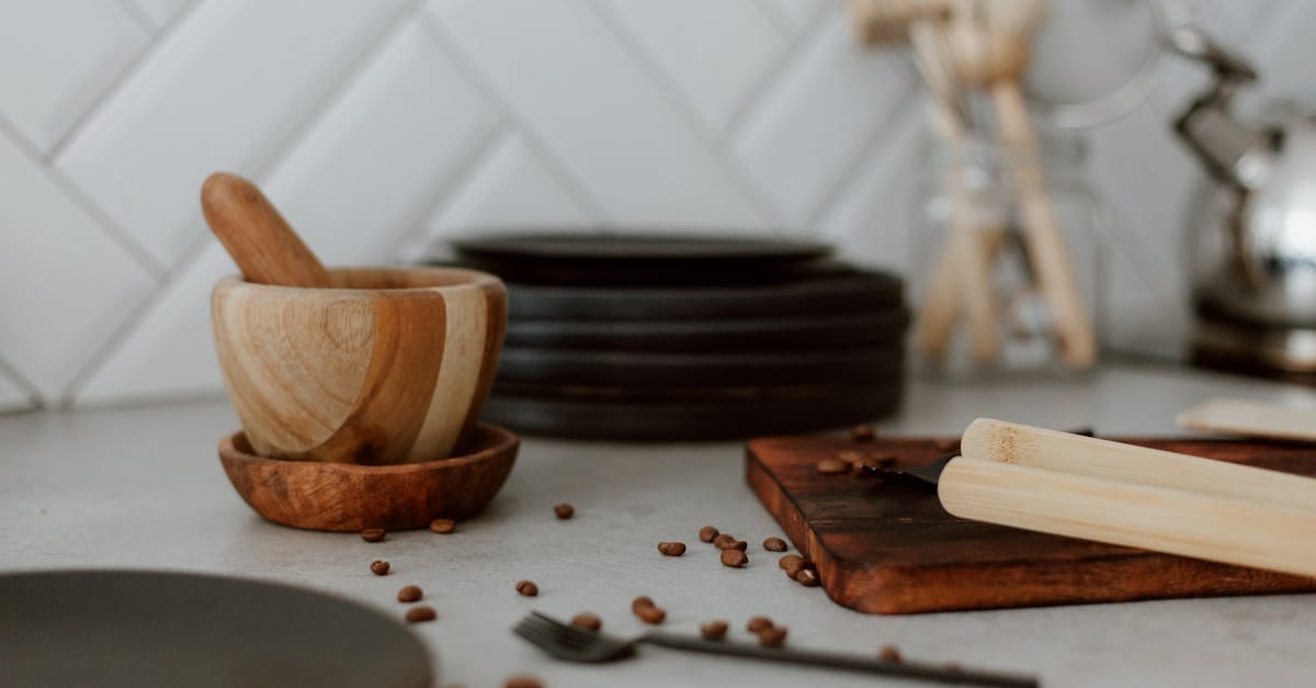 A wooden mortar and pestle, coffee beans, black plates, and utensils are arranged on a kitchen countertop with a white tile backsplash.
