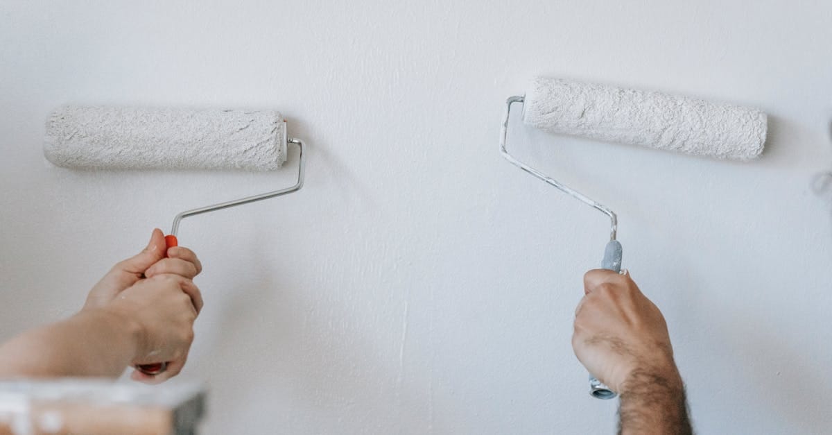 Two people using paint rollers to apply white paint to a wall.