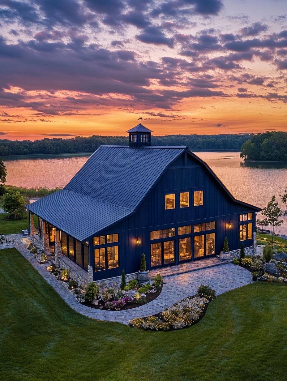 A large modern barn with many windows is lit from within at sunset, situated by a lake with a landscaped garden and manicured lawn.