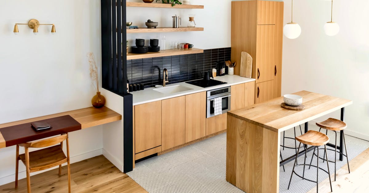 Modern kitchen with light wood cabinets, black backsplash, open shelves, island with stools, built-in desk area, and minimal decor under pendant lights.