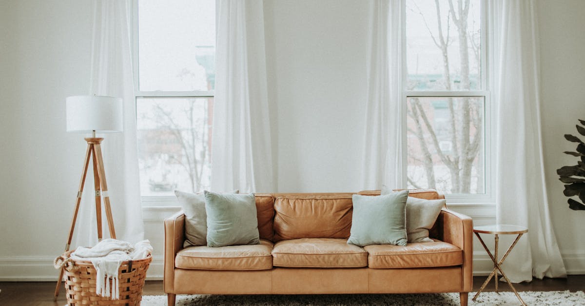 A tan leather sofa with four pillows sits between two large windows with white curtains; a floor lamp and woven basket are on the left, a small round table is on the right.