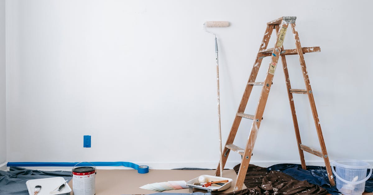 A wooden ladder, paint roller, open paint can, and painting supplies are set up in a room with a freshly painted white wall.