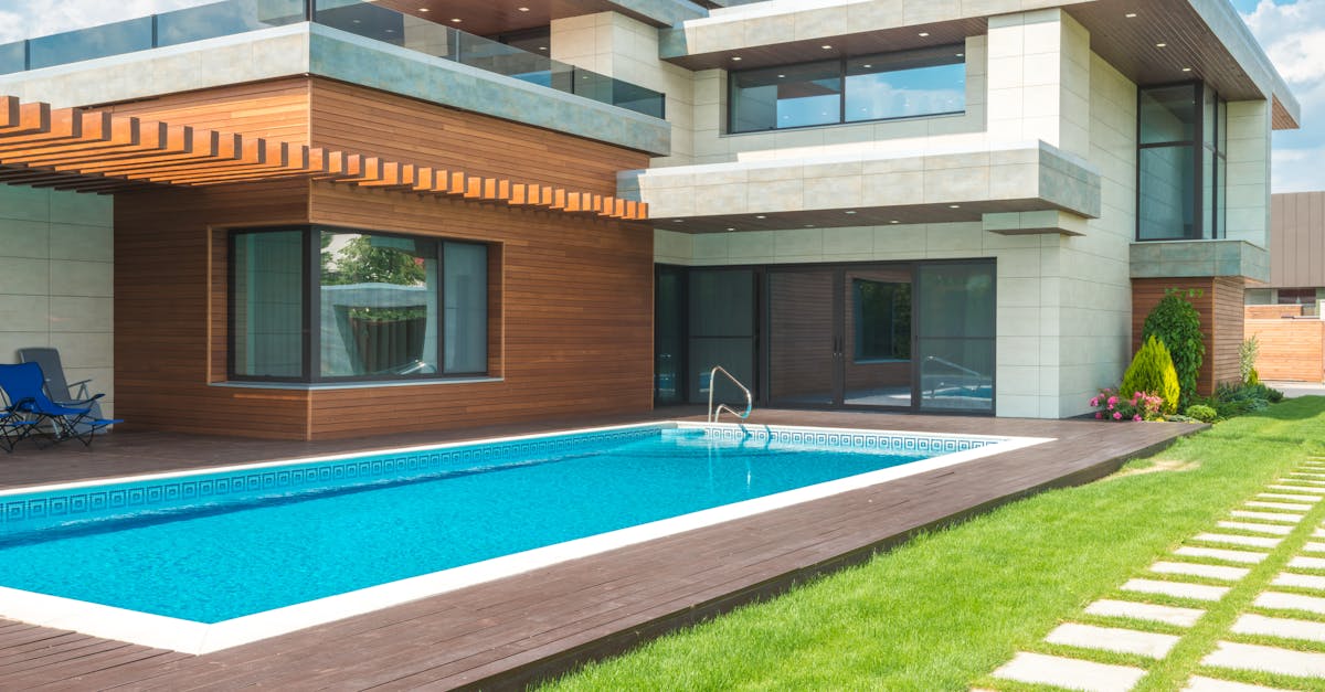 Modern two-story house with large windows, wooden and concrete exterior, and a rectangular swimming pool in the backyard surrounded by grass and a stone path.