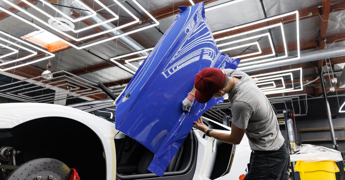 A person in a red cap applies a blue vinyl wrap to the door of a white car inside a workshop with geometric ceiling lights.