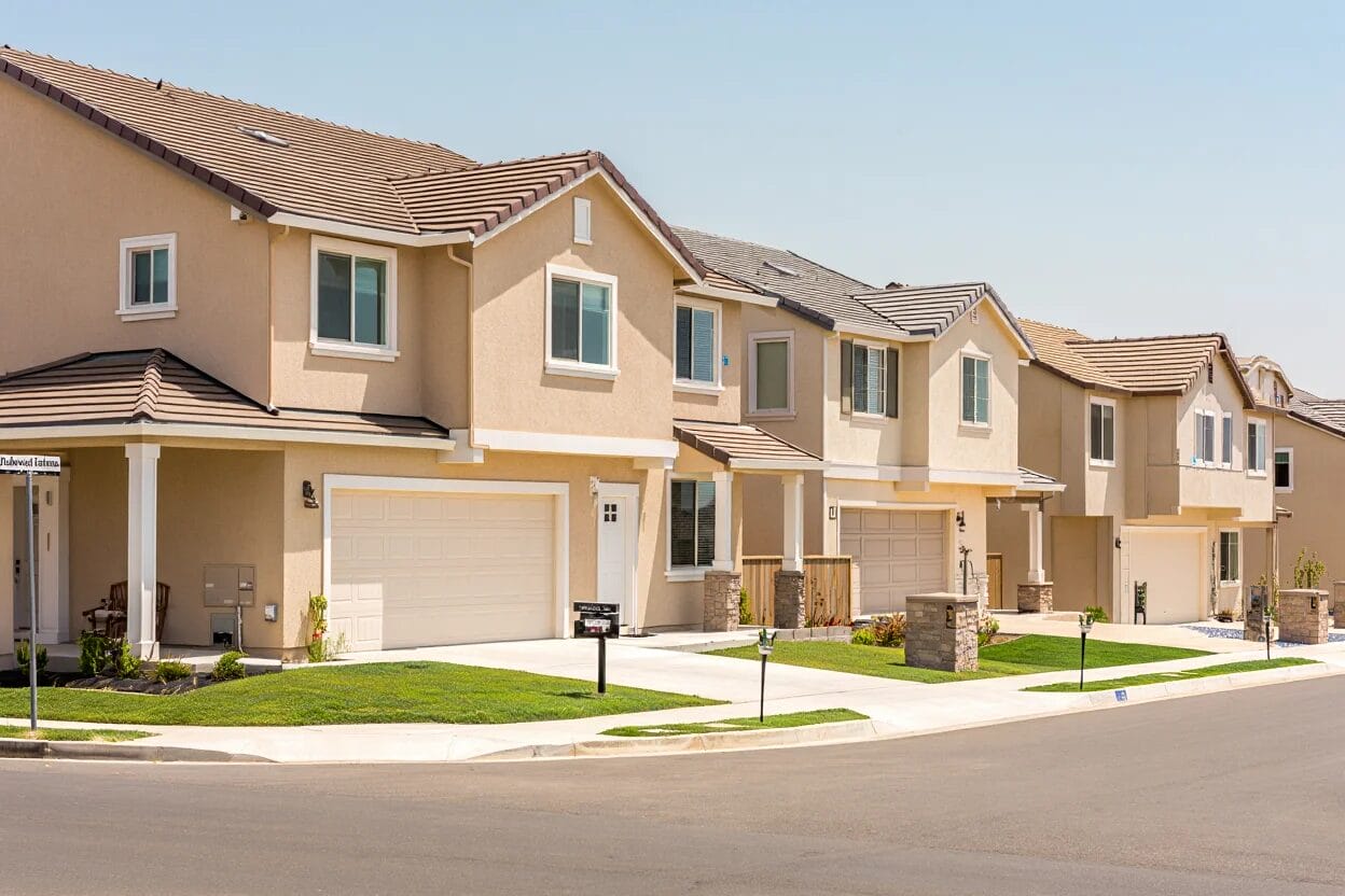 Row of modern two-story suburban houses with attached garages, manicured lawns, and a clean street under a clear sky.