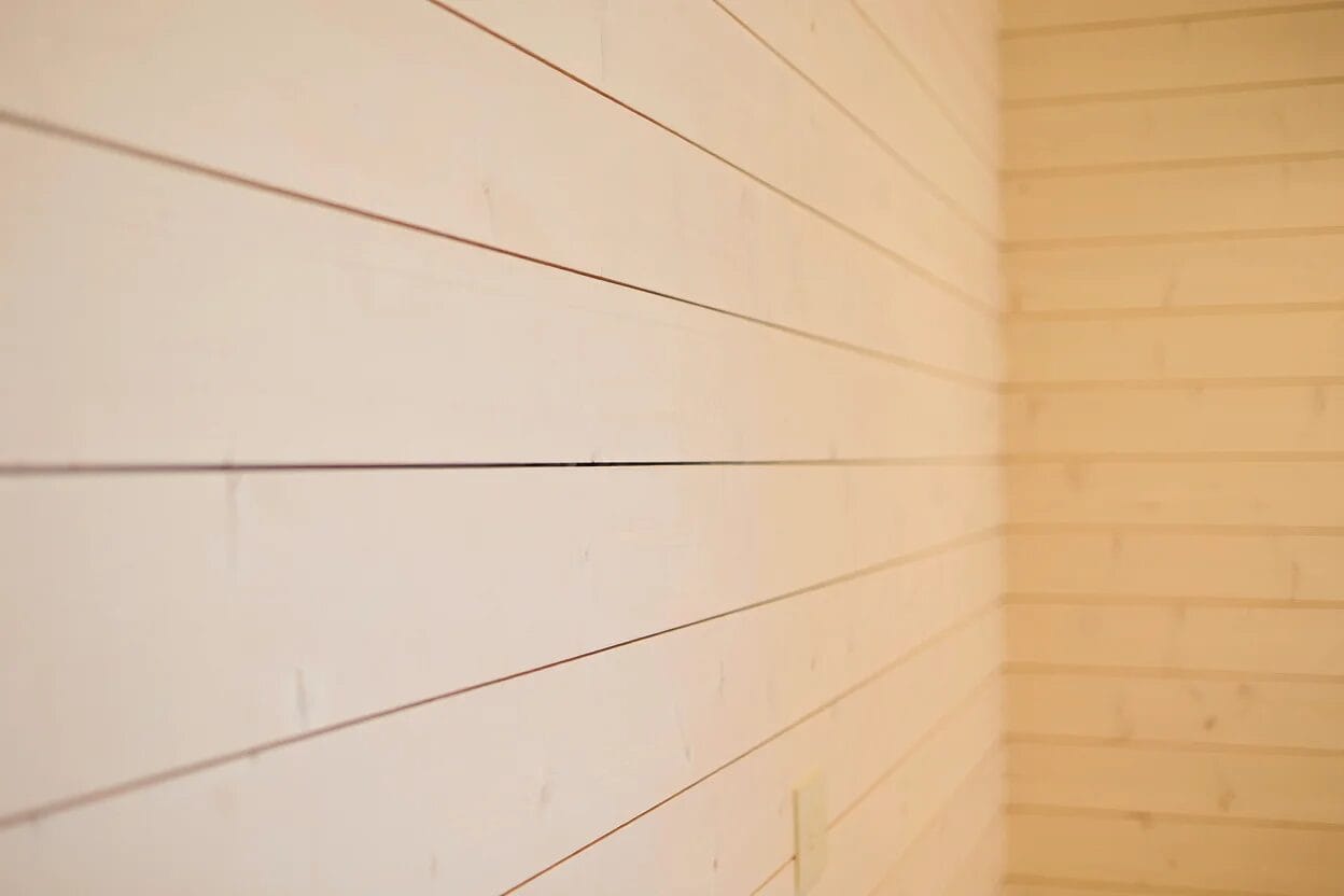 Close-up view of a light-colored wooden plank wall in a corner, with horizontal lines and a power outlet visible near the bottom.