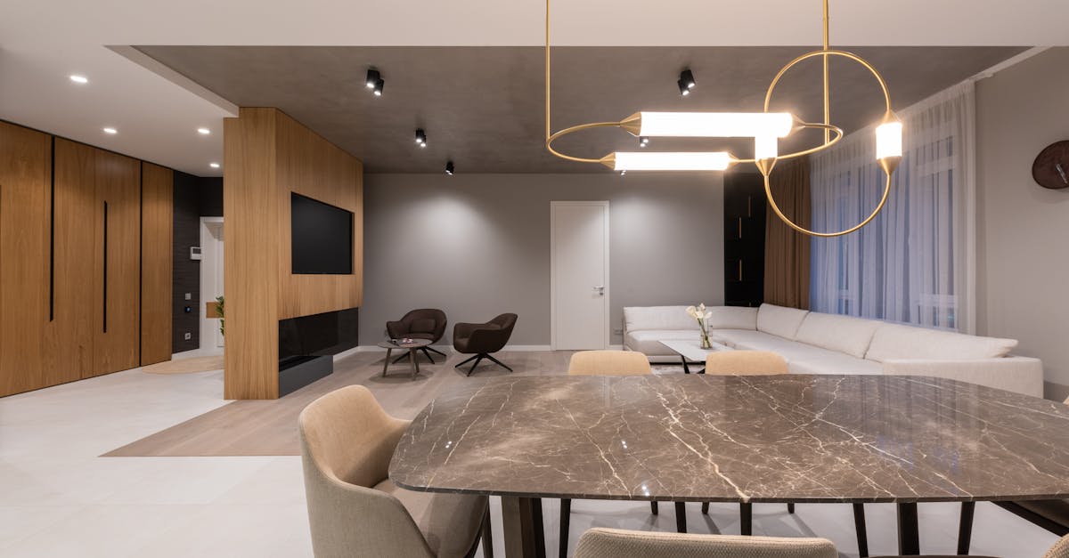 Modern open-plan living and dining area with a marble dining table, beige chairs, white sectional sofa, wooden accents, and contemporary lighting fixtures.