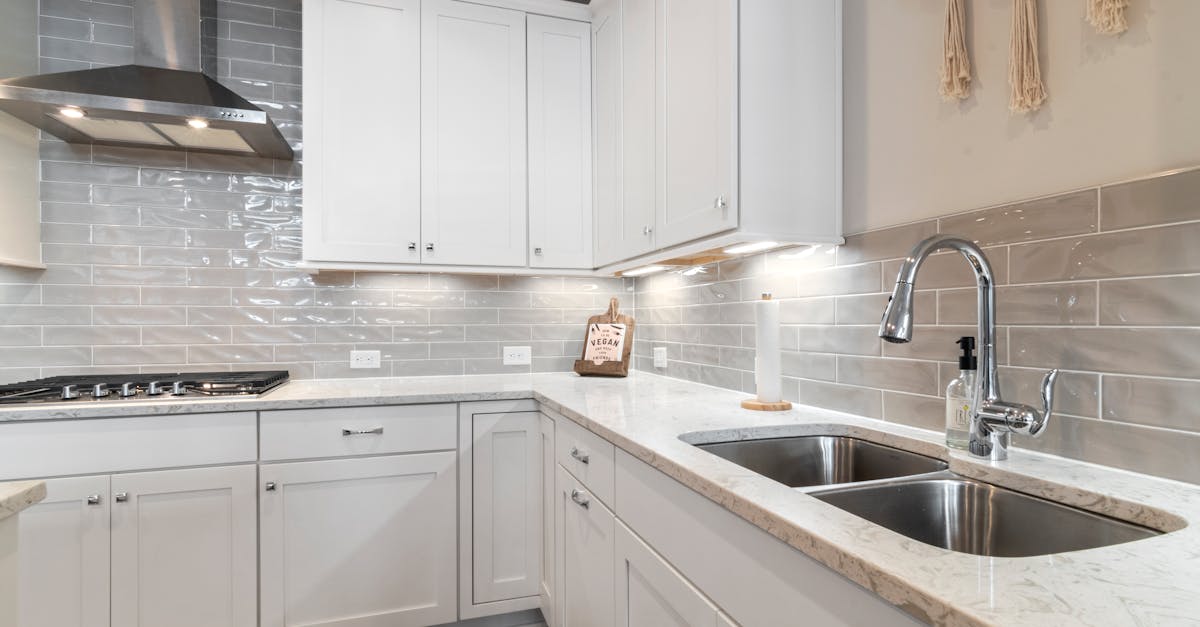 Modern kitchen with white cabinets, stainless steel appliances, double sink, marble countertop, and glossy gray subway tile backsplash. A paper towel holder and soap dispenser are near the sink.