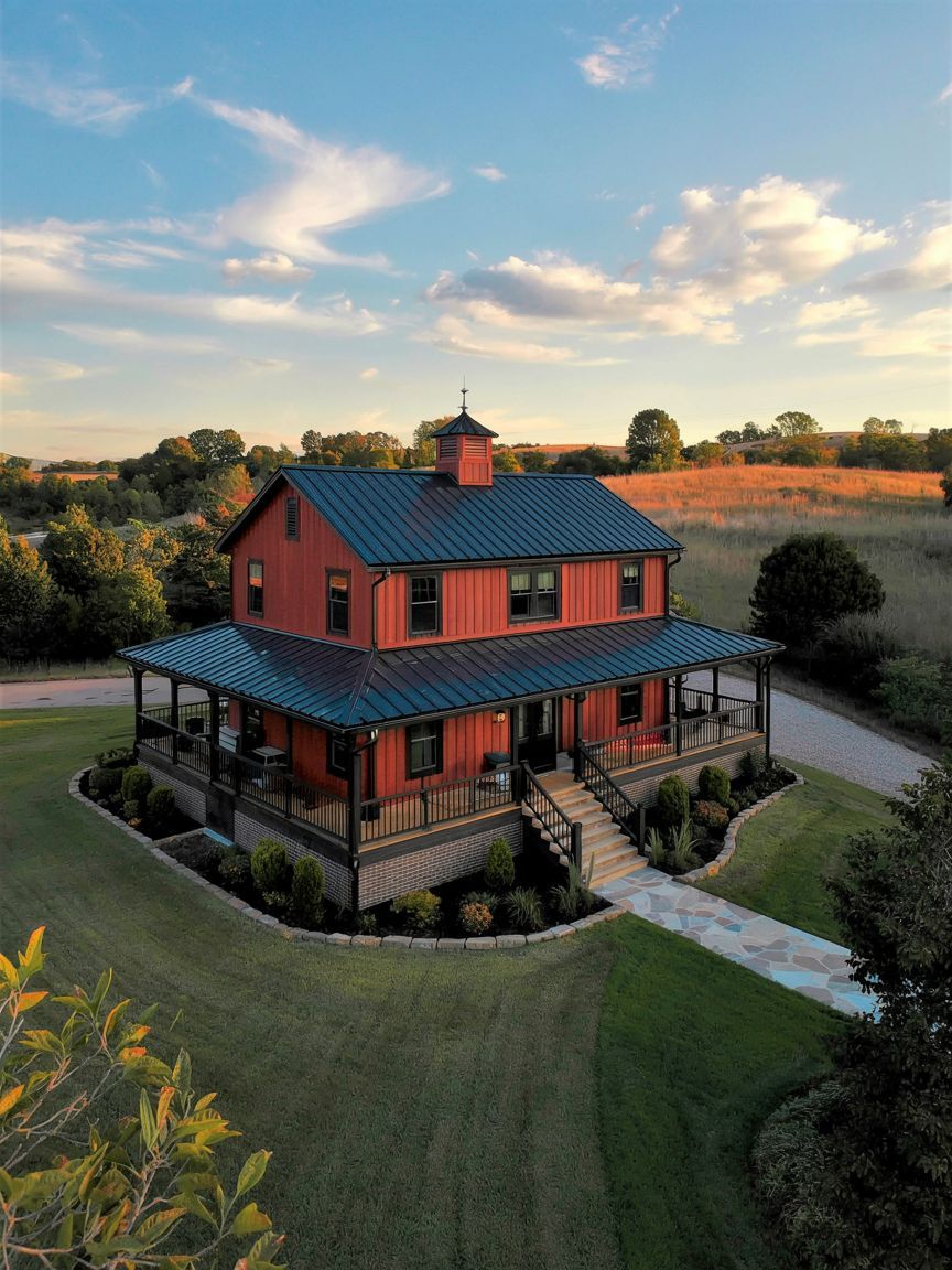 A red two-story farmhouse with a metal roof and wraparound porch sits in a landscaped yard surrounded by grass and trees, under a partly cloudy sky.