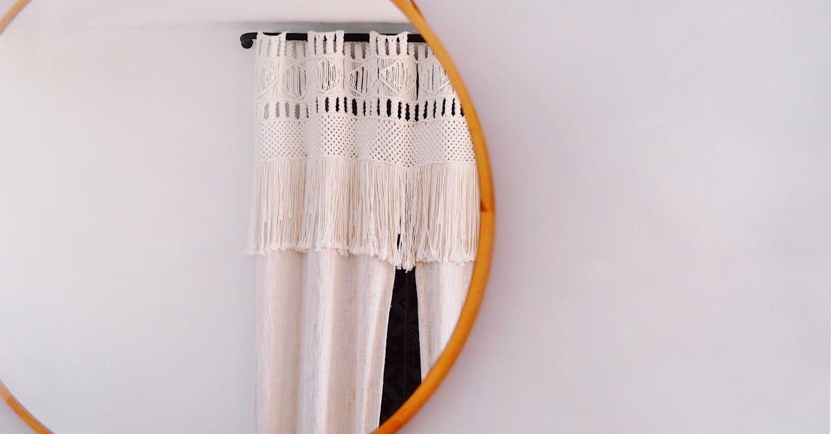 A round wooden-framed mirror reflects a white macrame curtain with fringe detailing hanging on a window.
