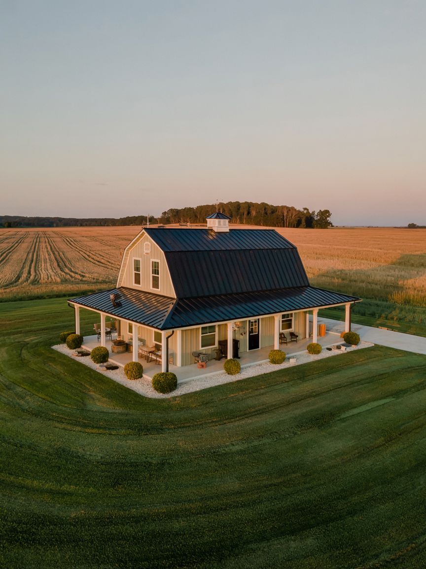 A farmhouse with a black roof and wraparound porch sits in the middle of a green lawn, surrounded by fields at sunset.