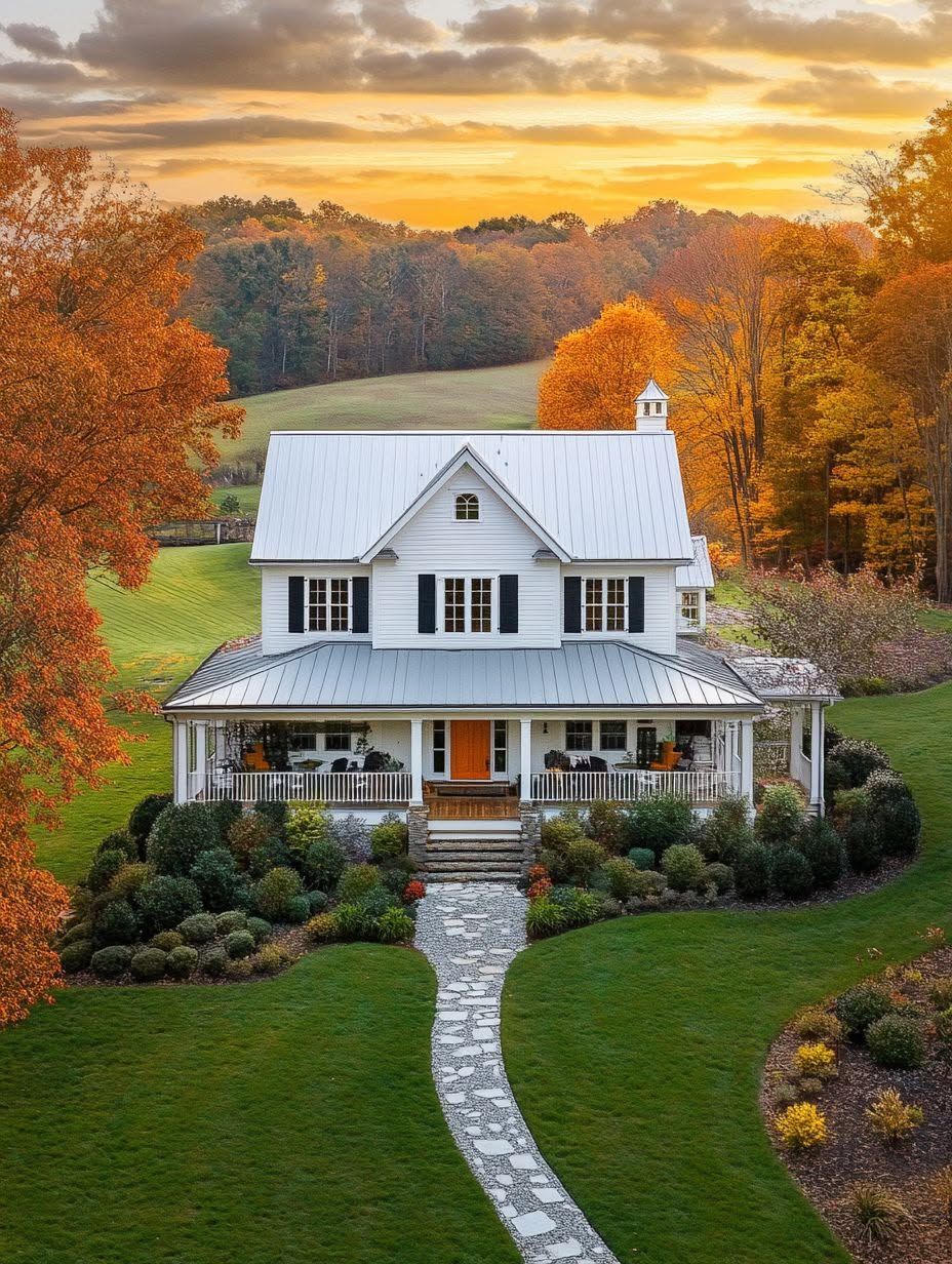 A white two-story house with a wraparound porch is surrounded by autumn trees and a well-kept lawn under a colorful sunset sky.