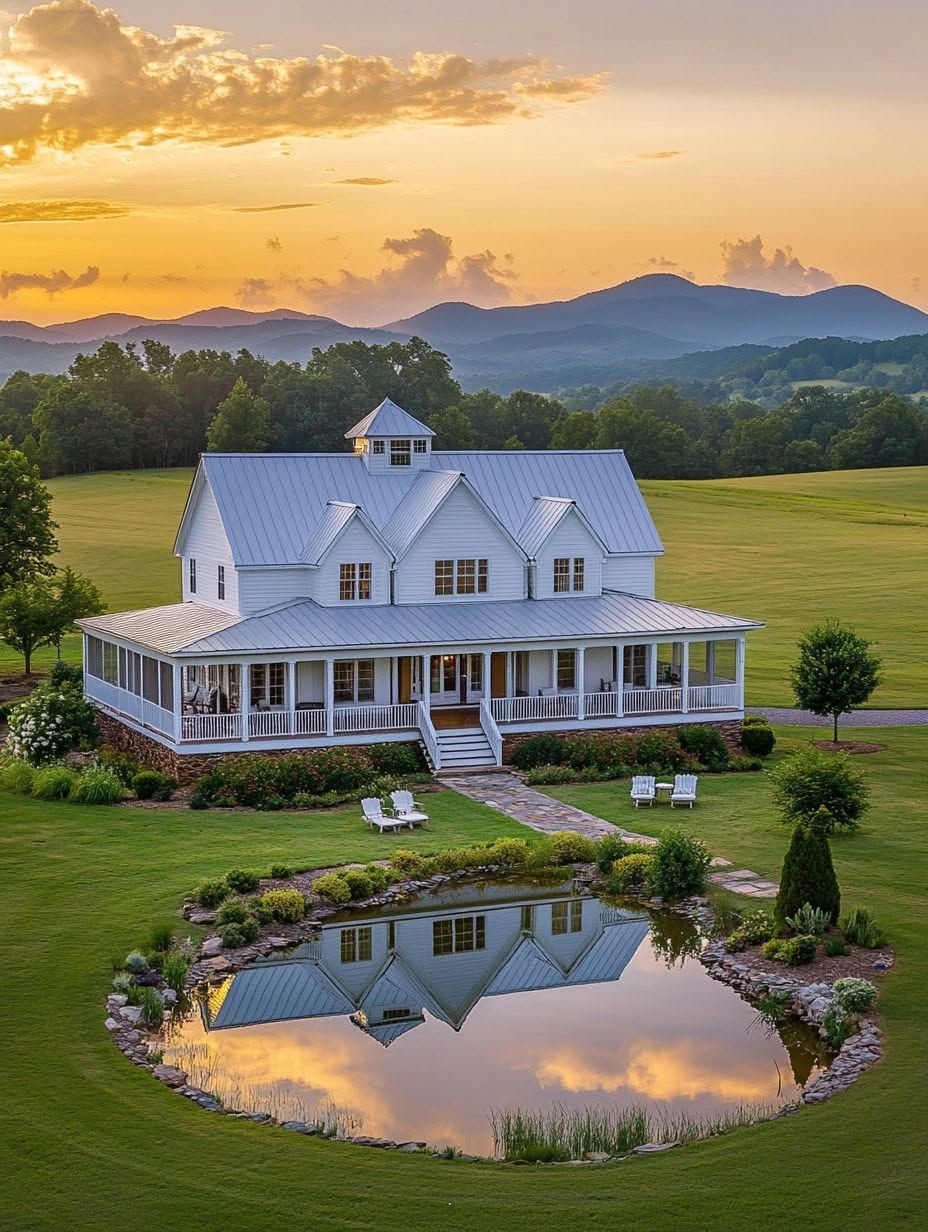 A large white farmhouse with a wraparound porch stands in a green field at sunset, reflected in a small pond in the foreground, with mountains in the distance.