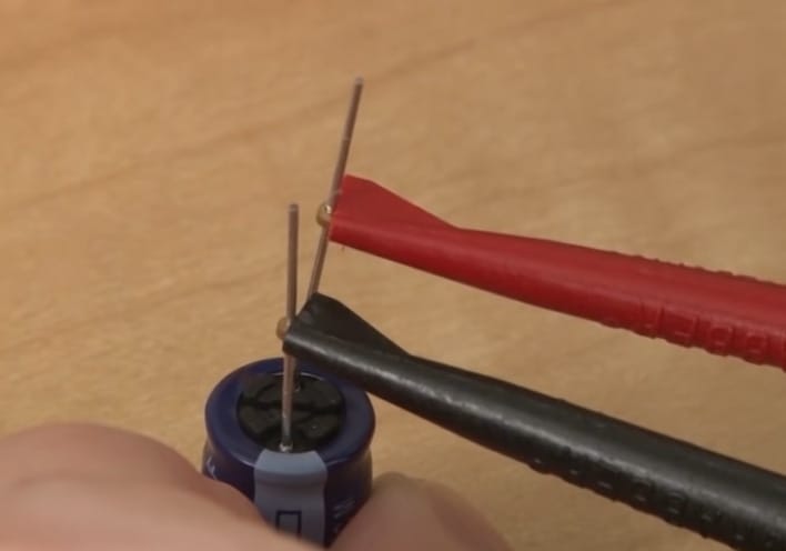 A person is using a pair of pliers to connect two wires in order to test a capacitor with a multimeter.