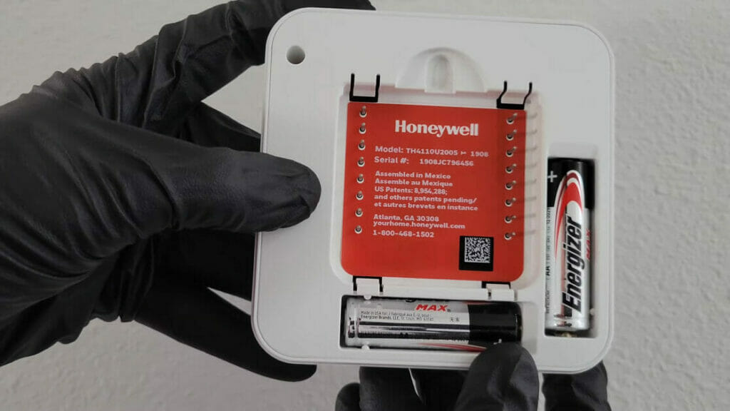 a hand wearing a black gloves holding a honeywell thermostat with energizer battery
