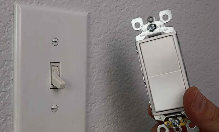 A man holding a new single pole light switch beside mounted old light switch on the wall