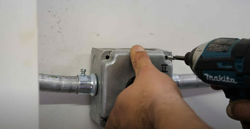A person securing the metal outlet box on the wall by drilling