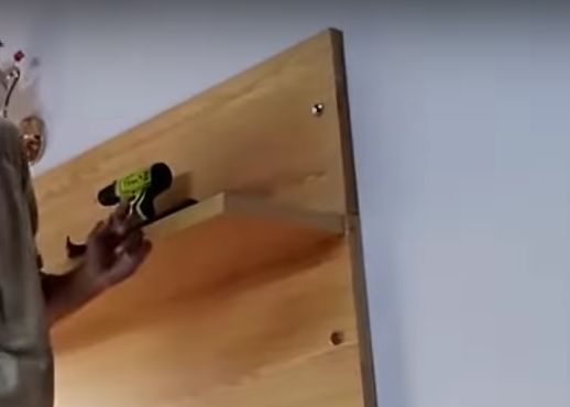 A man is using a drill to hide wires on a wooden shelf