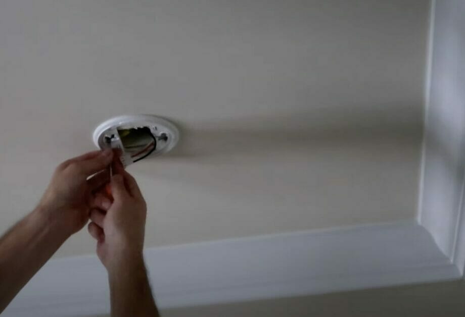 A person installing a base of a hard-wired smoke detector in a ceiling