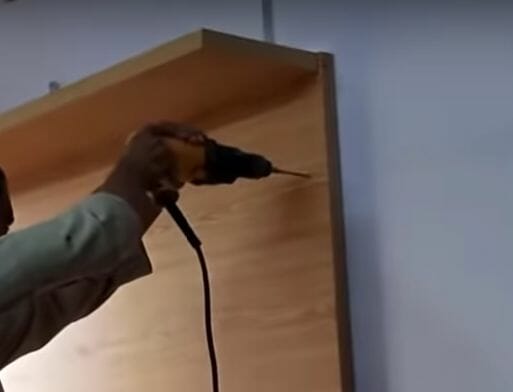 A man is using a drill to hide wires on a wooden tv shelf