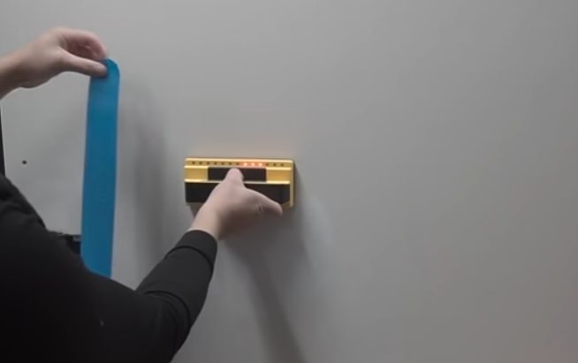 A person holding a blue tape and a stud finder placed at the wall