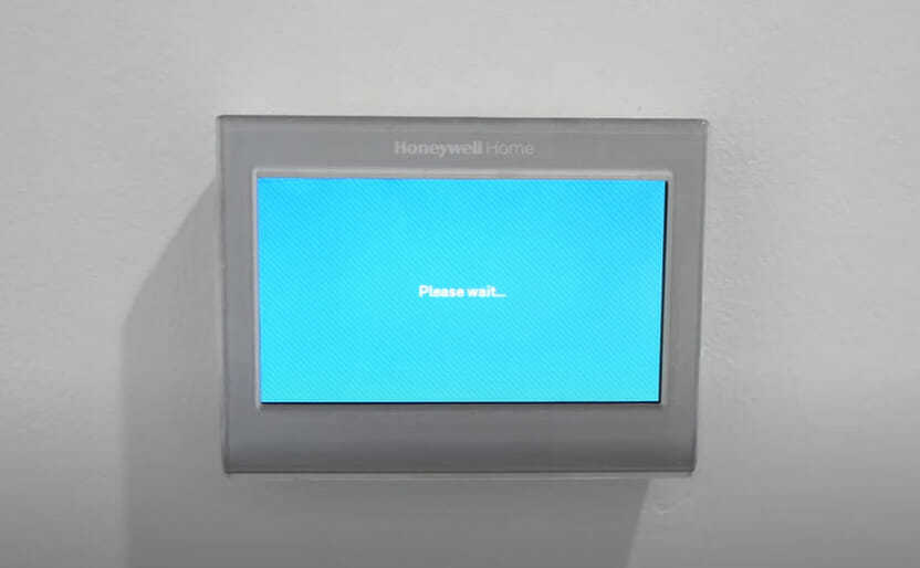 A Honeywell thermostat mounted on wall with message "Please wait..." on the screen