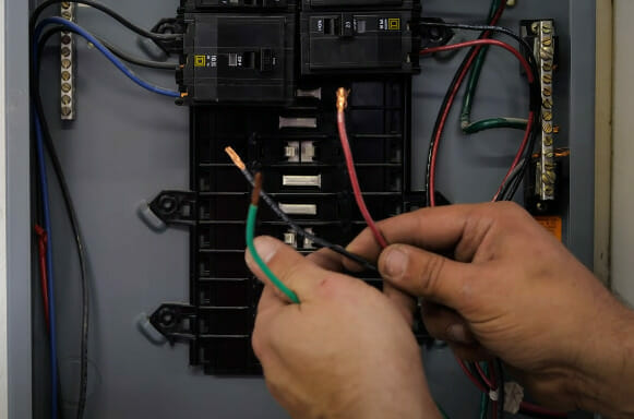 A person holding a green, black, and red wire in front of a main electrical panel