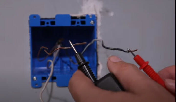 A person testing the wires in a blue electrical box using a multimeter