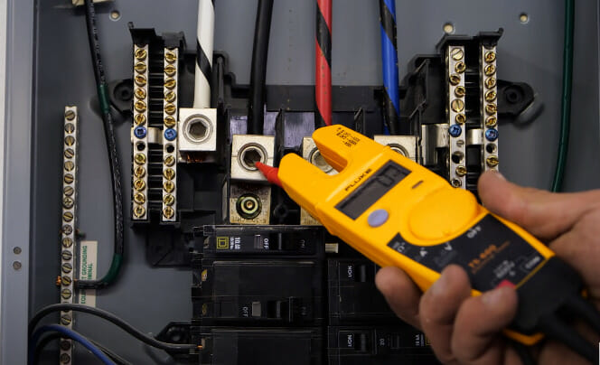 A person holding a yellow voltage tester in front of an electrical panel
