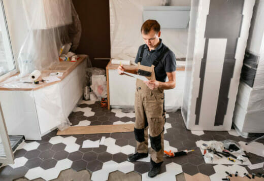 A man in overalls standing in front of a tiled kitchen holding and looking at a paper