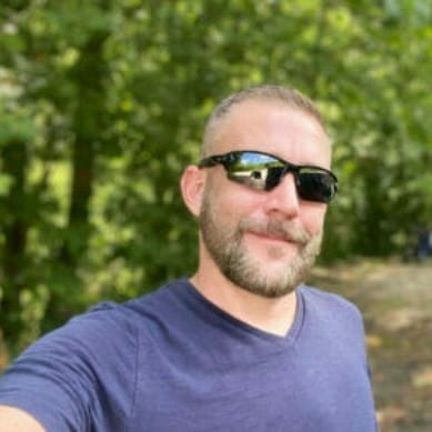 A man with a beard and sunglasses taking a selfie in the woods to share about himself.