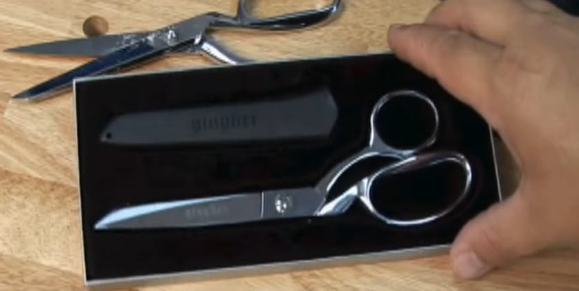 A person holding a scissors and shear on a box and seating besides it is another scissor